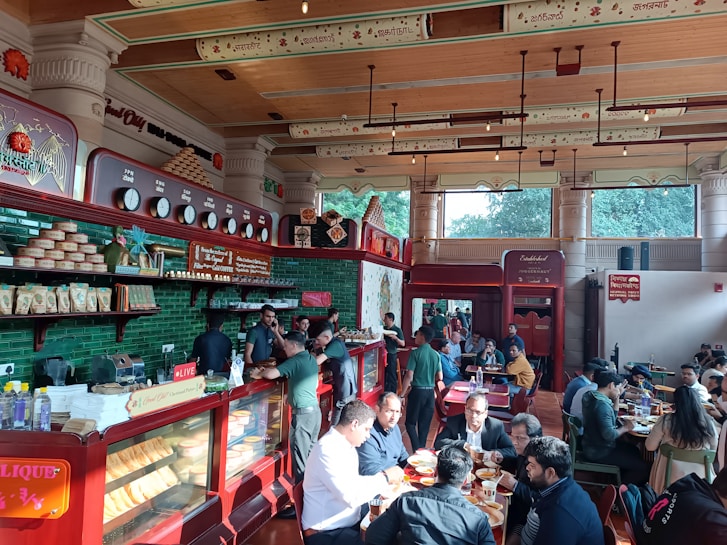 A bustling cafe interior with people sitting at tables, engaged in conversations and enjoying their meals. The decor features green tiles, wooden elements, and traditional designs on the walls and ceiling, creating a warm and inviting atmosphere. A display case filled with bakery items and a counter where staff are serving customers are visible on the left side.