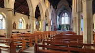 A spacious church interior featuring wooden pews aligned in rows, tall stone columns, and arched ceilings. Stained glass windows adorn the walls, letting colored light filter through. The altar is visible at the far end, with religious statues and decor throughout the space.