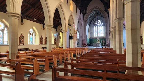 A spacious church interior featuring wooden pews aligned in rows, tall stone columns, and arched ceilings. Stained glass windows adorn the walls, letting colored light filter through. The altar is visible at the far end, with religious statues and decor throughout the space.