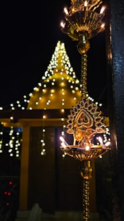 A beautifully ornate brass lamp is lit with multiple small flames, standing against the backdrop of a festively lit structure. Strings of golden lights form a conical shape, suggesting celebratory or festive ambiance.