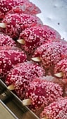 Several red confectionery treats are arranged in a tray, covered in small pink and white sugar balls. They are placed in decorative paper cups with floral patterns.