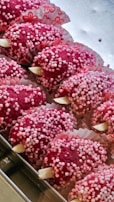 Several red confectionery treats are arranged in a tray, covered in small pink and white sugar balls. They are placed in decorative paper cups with floral patterns.