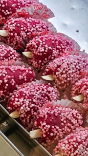 Several red confectionery treats are arranged in a tray, covered in small pink and white sugar balls. They are placed in decorative paper cups with floral patterns.