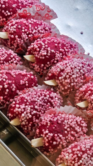 Several red confectionery treats are arranged in a tray, covered in small pink and white sugar balls. They are placed in decorative paper cups with floral patterns.