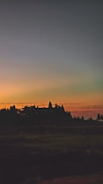 A landscape image featuring a silhouetted temple-like structure against a vibrant sunset. The sky transitions from deep blue at the top to warm orange near the horizon, and the structure is surrounded by darkened foliage and distant trees.