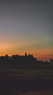 A landscape image featuring a silhouetted temple-like structure against a vibrant sunset. The sky transitions from deep blue at the top to warm orange near the horizon, and the structure is surrounded by darkened foliage and distant trees.
