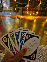 A hand holding a set of colorful cards with numbers and symbols, set in an indoor environment decorated with warm yellow string lights and a blurred background of various objects.