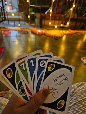A hand holding a set of colorful cards with numbers and symbols, set in an indoor environment decorated with warm yellow string lights and a blurred background of various objects.