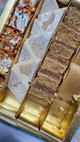 An assortment of Indian sweets presented in a box, featuring different types including those topped with silver foil. The sweets are divided into sections, showcasing a variety of colors and textures.