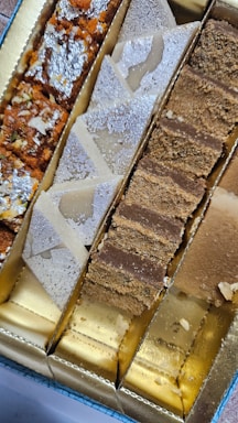 An assortment of Indian sweets presented in a box, featuring different types including those topped with silver foil. The sweets are divided into sections, showcasing a variety of colors and textures.