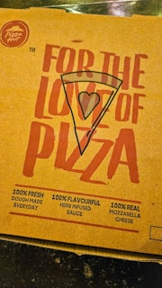 A pizza box lid with bold red text featuring the words 'For the love of pizza' and an illustration of a pizza slice with a heart symbol in the center. It includes promotional claims such as 100% fresh dough made every day, 100% flavorful herb-infused sauce, and 100% real mozzarella cheese. The Pizza Hut logo is visible in the top left corner.