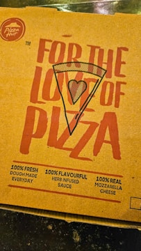 A pizza box lid with bold red text featuring the words 'For the love of pizza' and an illustration of a pizza slice with a heart symbol in the center. It includes promotional claims such as 100% fresh dough made every day, 100% flavorful herb-infused sauce, and 100% real mozzarella cheese. The Pizza Hut logo is visible in the top left corner.