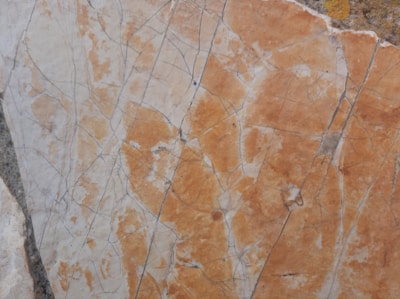 A close-up of a marble surface featuring intricate cracks and veining patterns in shades of orange, beige, and light gray. The texture appears smooth with natural imperfections, creating a visually interesting and organic design.