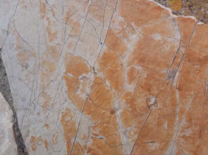 A close-up of a marble surface featuring intricate cracks and veining patterns in shades of orange, beige, and light gray. The texture appears smooth with natural imperfections, creating a visually interesting and organic design.