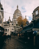 A cobblestone street in a European city is lined with cafes and shops, leading towards a large basilica with domed roofs and intricate architectural details. People walk along the street, and one side features a tree with autumn foliage. The sky is partly cloudy, adding a moody atmosphere to the scene.