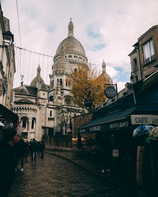 A cobblestone street in a European city is lined with cafes and shops, leading towards a large basilica with domed roofs and intricate architectural details. People walk along the street, and one side features a tree with autumn foliage. The sky is partly cloudy, adding a moody atmosphere to the scene.