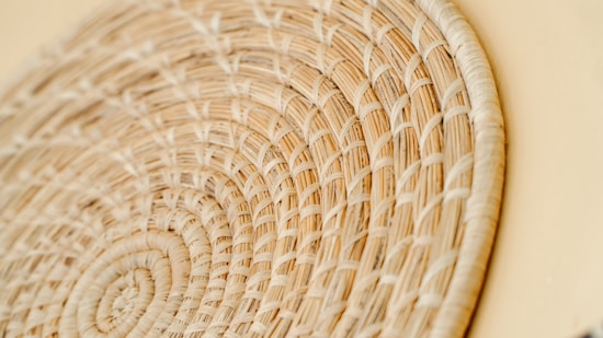 A close-up of a woven wicker basket with intricate patterns, showcasing the texture and craftsmanship of the intertwined natural fibers. The basket appears to have a circular design with a light beige color.