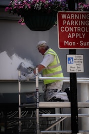 A person in a high-visibility vest is painting a wall while kneeling on a scaffold. Above, there is a hanging basket with purple and pink flowers. Nearby, two signs provide parking control information and goods vehicle loading instructions.