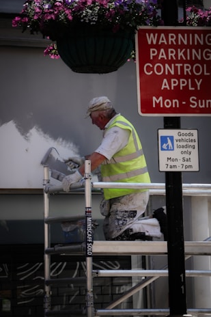 A person in a high-visibility vest is painting a wall while kneeling on a scaffold. Above, there is a hanging basket with purple and pink flowers. Nearby, two signs provide parking control information and goods vehicle loading instructions.