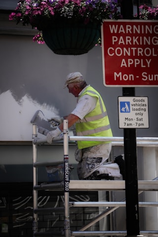 A person in a high-visibility vest is painting a wall while kneeling on a scaffold. Above, there is a hanging basket with purple and pink flowers. Nearby, two signs provide parking control information and goods vehicle loading instructions.