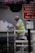 A person in a high-visibility vest is painting a wall while kneeling on a scaffold. Above, there is a hanging basket with purple and pink flowers. Nearby, two signs provide parking control information and goods vehicle loading instructions.