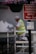 A person in a high-visibility vest is painting a wall while kneeling on a scaffold. Above, there is a hanging basket with purple and pink flowers. Nearby, two signs provide parking control information and goods vehicle loading instructions.