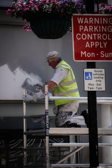 A person in a high-visibility vest is painting a wall while kneeling on a scaffold. Above, there is a hanging basket with purple and pink flowers. Nearby, two signs provide parking control information and goods vehicle loading instructions.