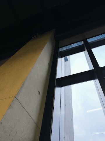 A modern architectural angle showing a concrete wall with a yellow section and large glass windows that extend upwards.