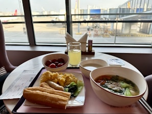 A table by a window at an airport lounge features a breakfast spread including scrambled eggs, an Asian-style soup with vegetables, bread, a bowl of cherry tomatoes, and a glass of juice. A boarding pass is placed on the table.
