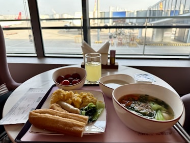 A table by a window at an airport lounge features a breakfast spread including scrambled eggs, an Asian-style soup with vegetables, bread, a bowl of cherry tomatoes, and a glass of juice. A boarding pass is placed on the table.