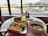 A table by a window at an airport lounge features a breakfast spread including scrambled eggs, an Asian-style soup with vegetables, bread, a bowl of cherry tomatoes, and a glass of juice. A boarding pass is placed on the table.