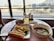 A table by a window at an airport lounge features a breakfast spread including scrambled eggs, an Asian-style soup with vegetables, bread, a bowl of cherry tomatoes, and a glass of juice. A boarding pass is placed on the table.
