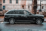 A luxurious black SUV is parked on a city street, adjacent to a black metal fence and large tree. The vehicle features prominent, stylish wheels and a sleek exterior design. The background includes a historic building with large windows visible behind the fence.