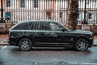 A luxurious black SUV is parked on a city street, adjacent to a black metal fence and large tree. The vehicle features prominent, stylish wheels and a sleek exterior design. The background includes a historic building with large windows visible behind the fence.