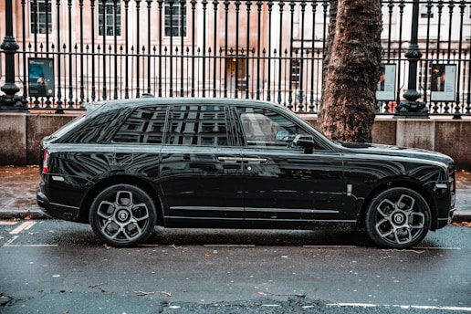 A luxurious black SUV is parked on a city street, adjacent to a black metal fence and large tree. The vehicle features prominent, stylish wheels and a sleek exterior design. The background includes a historic building with large windows visible behind the fence.