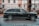 A luxurious black SUV is parked on a city street, adjacent to a black metal fence and large tree. The vehicle features prominent, stylish wheels and a sleek exterior design. The background includes a historic building with large windows visible behind the fence.