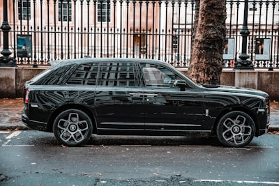 A luxurious black SUV is parked on a city street, adjacent to a black metal fence and large tree. The vehicle features prominent, stylish wheels and a sleek exterior design. The background includes a historic building with large windows visible behind the fence.
