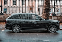 A luxurious black SUV is parked on a city street, adjacent to a black metal fence and large tree. The vehicle features prominent, stylish wheels and a sleek exterior design. The background includes a historic building with large windows visible behind the fence.