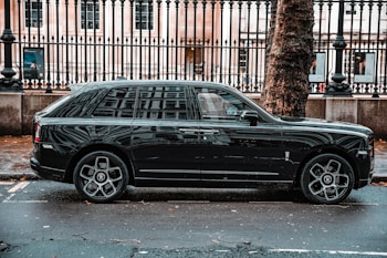 A luxurious black SUV is parked on a city street, adjacent to a black metal fence and large tree. The vehicle features prominent, stylish wheels and a sleek exterior design. The background includes a historic building with large windows visible behind the fence.
