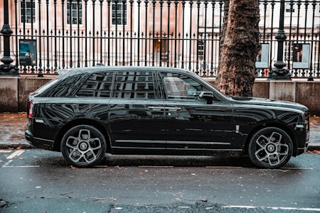 A luxurious black SUV is parked on a city street, adjacent to a black metal fence and large tree. The vehicle features prominent, stylish wheels and a sleek exterior design. The background includes a historic building with large windows visible behind the fence.