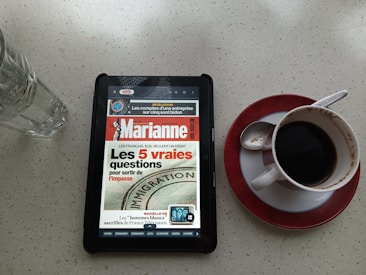 A tablet displaying a French magazine cover is placed on a light-colored speckled table surface. Next to the tablet, there's a white cup of black coffee on a red-bordered saucer with a spoon resting inside. A tall empty glass is positioned to the left of the tablet.