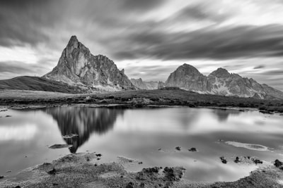 A dramatic black and white landscape featuring rugged mountains reflected in a calm body of water. The clouds appear blurred, implying long exposure photography, adding a dynamic quality to the serene scene.