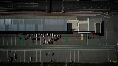 An aerial view of a large parking lot with several parked cars near long commercial buildings. The lot is mostly empty, with lines marking parking spaces and green lanes possibly indicating pedestrian paths or bike lanes. Surrounding the lot are wide roads with multiple lanes.