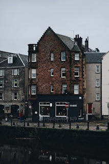 A quaint, old building with red brickwork stands prominently by a canal. The structure features multiple windows in varying sizes and a dark, shingled roof. Below, a café named 'Toast' occupies the ground floor, with a small seating area outdoors. The surrounding area is quiet, with cobblestone paths and adjacent buildings showcasing similar architectural styles.