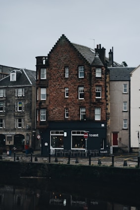 A quaint, old building with red brickwork stands prominently by a canal. The structure features multiple windows in varying sizes and a dark, shingled roof. Below, a café named 'Toast' occupies the ground floor, with a small seating area outdoors. The surrounding area is quiet, with cobblestone paths and adjacent buildings showcasing similar architectural styles.