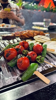 A close-up of a grill with skewers of roasted cherry tomatoes, green chili peppers, and onion slices. In the background, a person is using a smartphone while seated at a table. There are plates and glasses visible on the table, and the setting appears like an outdoor or restaurant environment with greenery and decorative lights.