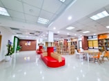 A modern and bright indoor space with high ceilings and a tiled floor. The room features a combination of red seating, wooden shelves filled with books, and colorful chairs around a table. On one side, there's a brick wall with a lit signage displaying 'CIE'. The space is well-lit with multiple ceiling lights and decorated with potted plants.