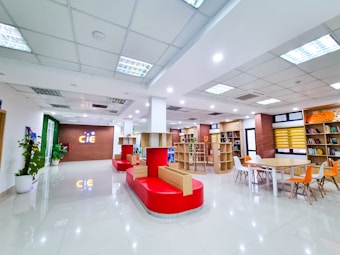 A modern and bright indoor space with high ceilings and a tiled floor. The room features a combination of red seating, wooden shelves filled with books, and colorful chairs around a table. On one side, there's a brick wall with a lit signage displaying 'CIE'. The space is well-lit with multiple ceiling lights and decorated with potted plants.
