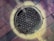 A manhole cover labeled 'Boston Edison Company Steam' is centered on a brick-paved surface. The cover features a pattern of hexagonal shapes, and there is a circular light or discoloration around it, giving an interesting visual effect.