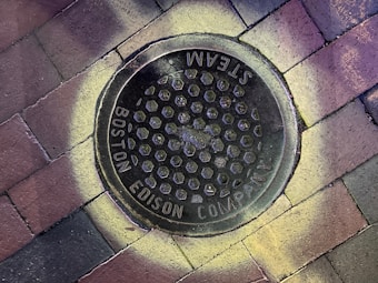 A manhole cover labeled 'Boston Edison Company Steam' is centered on a brick-paved surface. The cover features a pattern of hexagonal shapes, and there is a circular light or discoloration around it, giving an interesting visual effect.